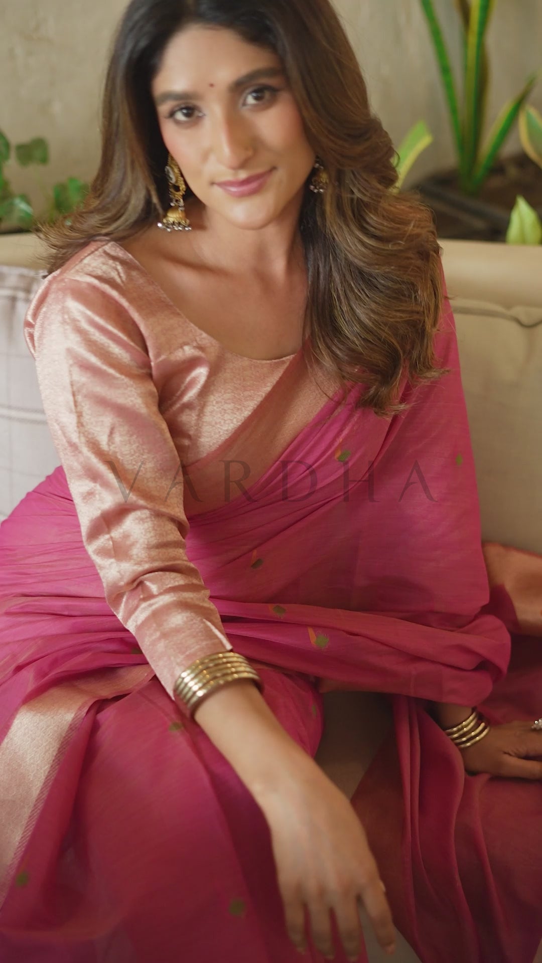 A woman wearing a traditional Indian saree with a metallic rose gold blouse and a magenta pink saree with gold zari embroidery, sitting on a beige sofa. Beautiful model sitting gracefully, showcasing the perfect drape, delicate body motifs, and vibrant magenta hue of the cotton jacquard festive saree.