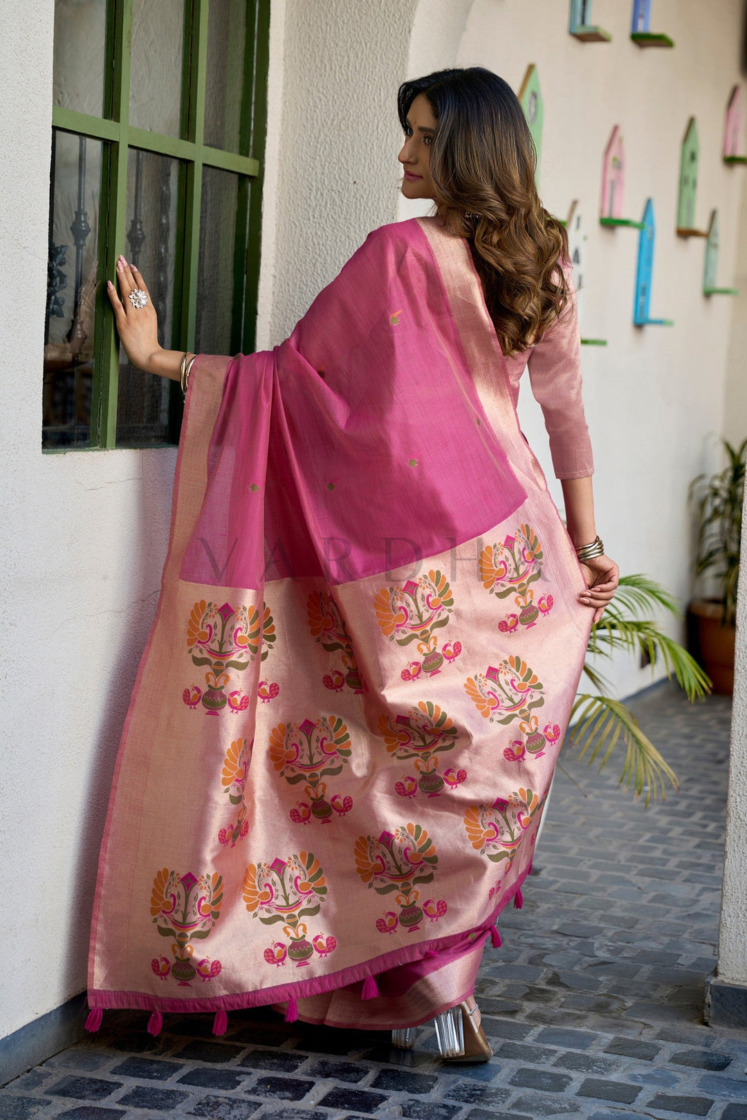 Woman wearing a pink and beige saree with floral patterns, standing against a wall. Back view of the Rani pink cotton jacquard saree displaying the grand oversized woven Paithani peacock motifs alongside matching pink fancy tassels on the designer pallu.
