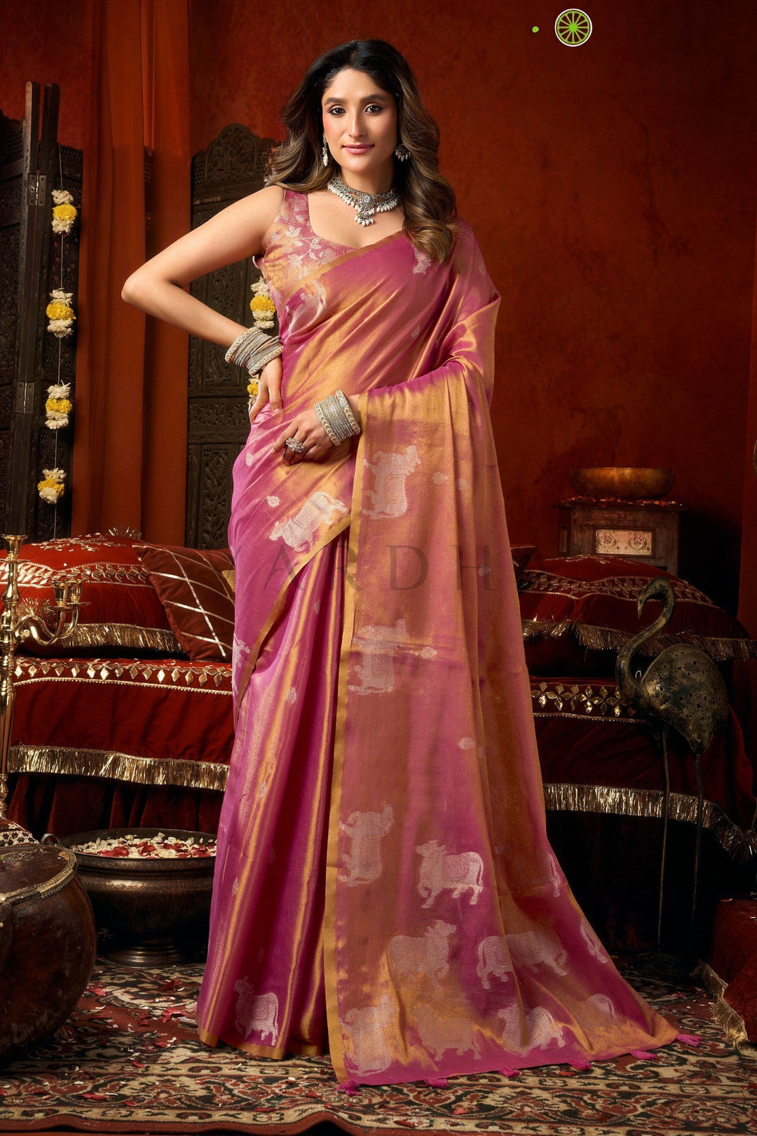 Woman in a pink and orange saree with animal patterns in an ornate room. Full front view of elegant pink heavy tissue saree featuring allover silver woven cow buttas and a rich golden zari border.