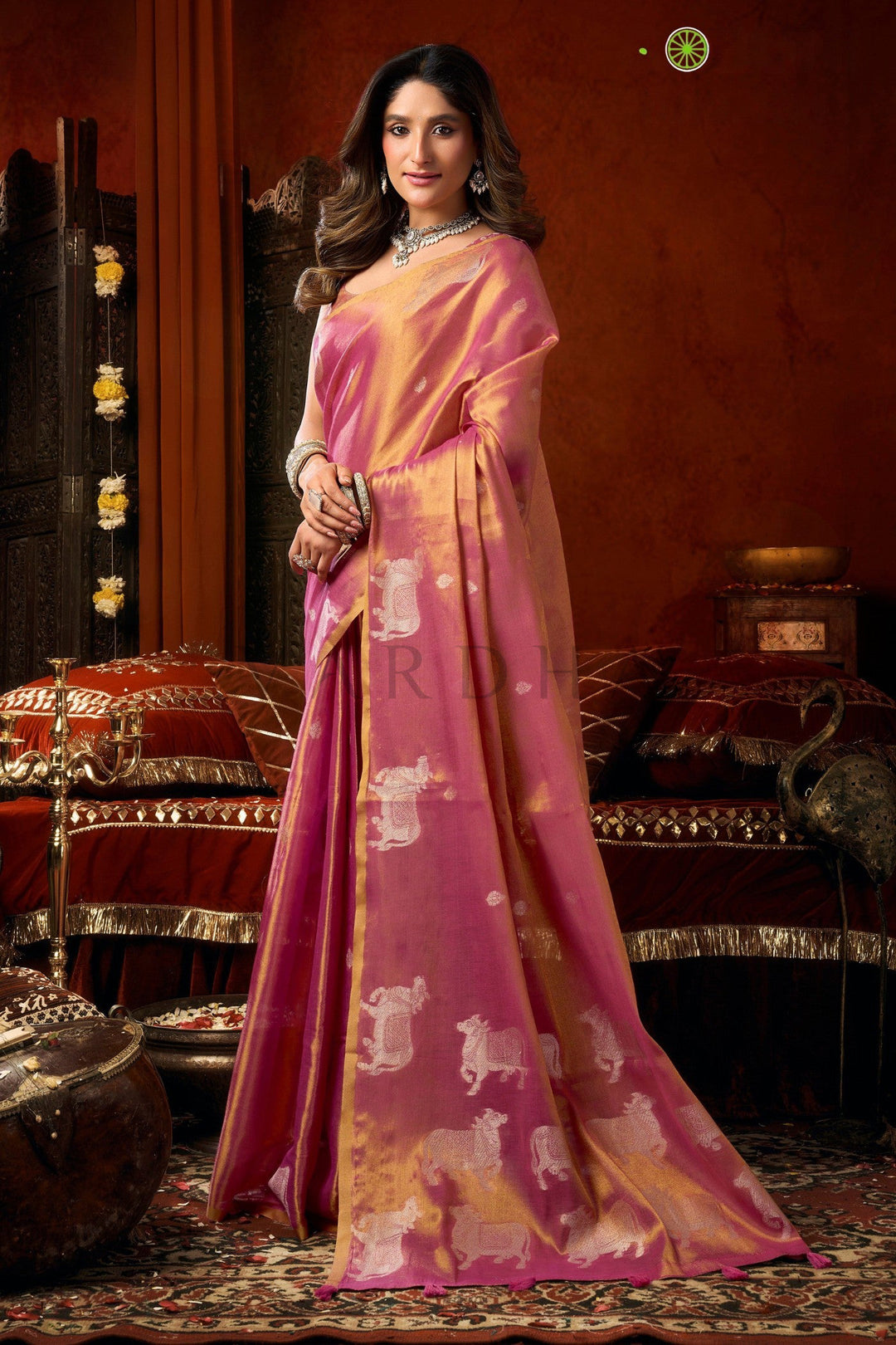 Woman wearing a pink saree with a pattern in an ornate room. Side profile of a woman gracefully draping a dual-tone magenta pink heavy tissue saree for wedding and festive occasions.