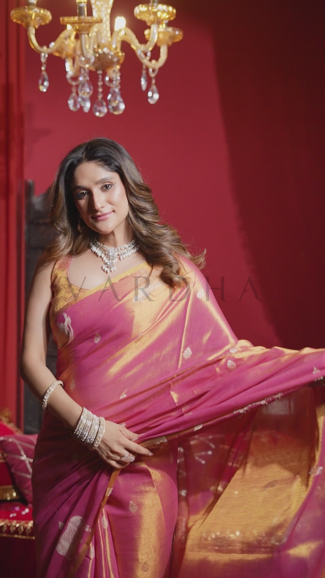 A woman wearing a pink and gold saree with silver jewelry, posing in front of a red wall with a gold chandelier. Dynamic flowing shot of the pink heavy tissue silk saree, emphasizing its radiant gold undertones and elegant drape for special events.