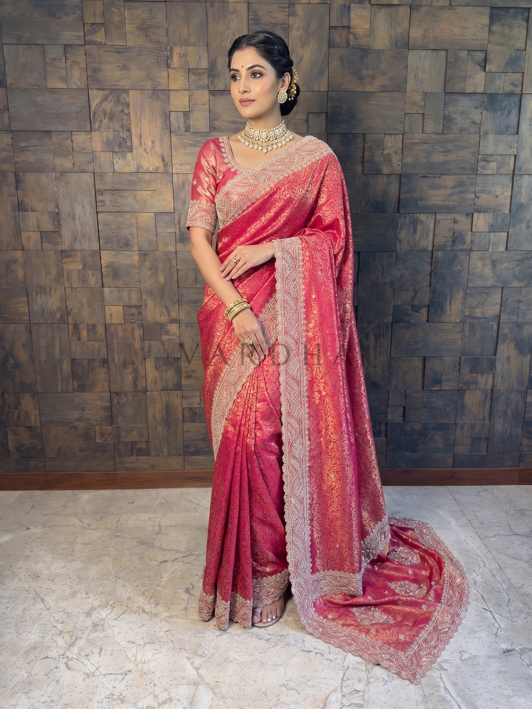Woman in a red saree with gold patterns standing against a textured wall.
