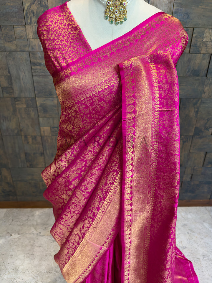 Pink saree with gold patterns on a mannequin against a wooden paneled wall. Close-up of House of Vardha Pink Kanjivaram Silk Saree with Zari Weave highlighting detailed gold zari floral patterns on the pleats and body. We offer Ready to wear, Pre drape service, Saree Add on, Custom Stitched Blouse, Fall and Edging, Gift Packaging at Complimentary Price!