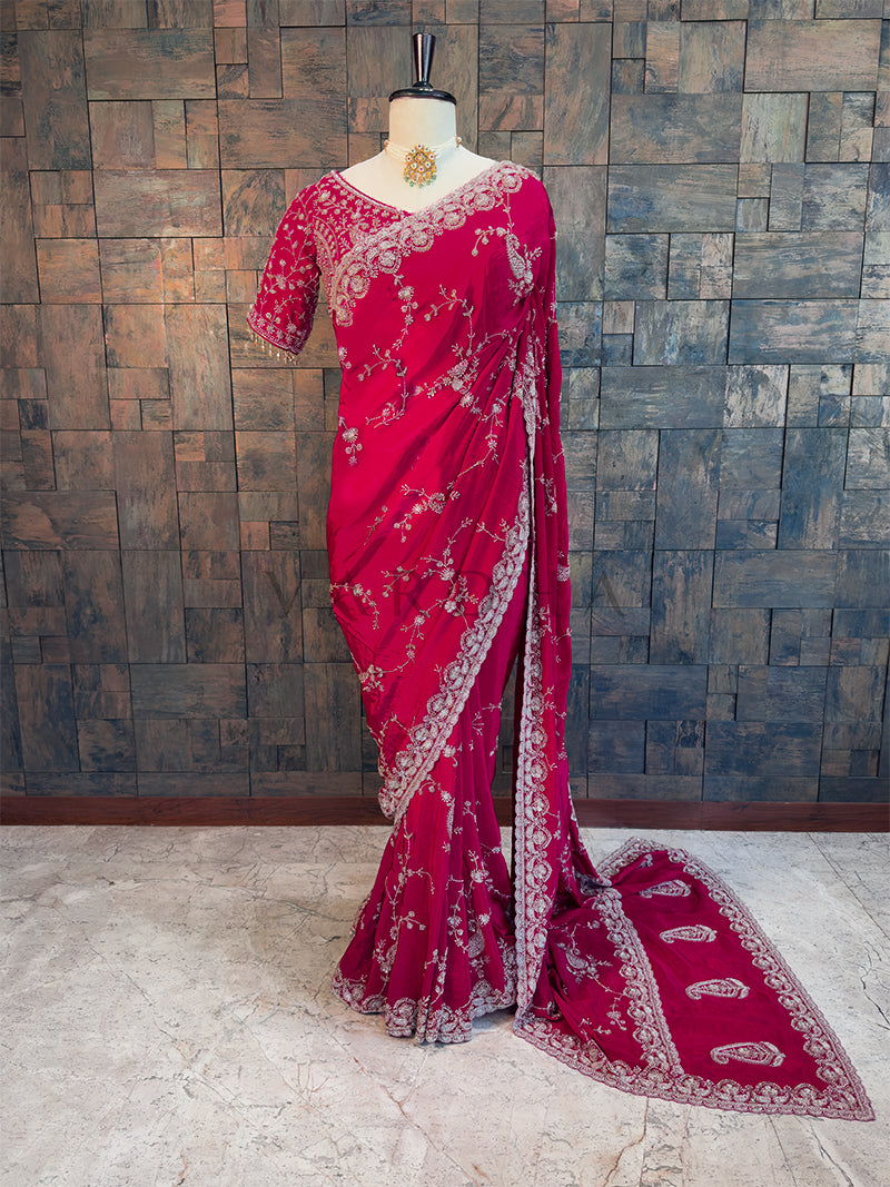 Pink saree with white patterns on a mannequin against a wooden paneled wall. Full View Pink Satin Crepe Handwork Saree for Sangeet. House of Vardha Pink Satin Crepe Saree featuring heavy silver embellishments and a matching scalloped border, elegantly draped on a mannequin. We offer Ready to wear, Pre drape service, Saree Add on, Custom Stitched Blouse, Fall and Edging, Gift Packaging at Complimentary Price!
