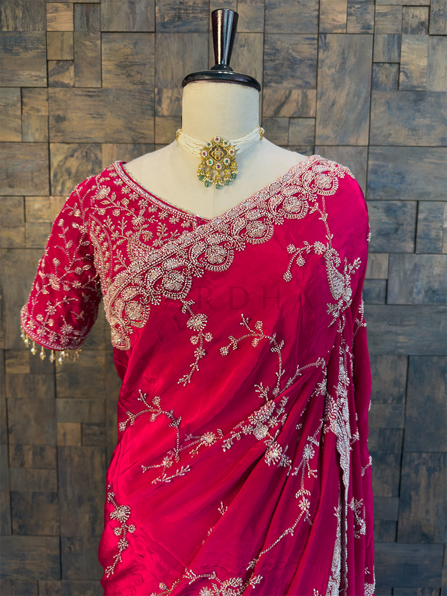 Pink saree with white embroidery on a mannequin against a wooden paneled wall. Designer Silver Embroidered Blouse paired with Pink Saree. Elegant Pink Satin Crepe Saree by House of Vardha showcasing exquisite all-over floral bead embroidery with a matching heavily embellished blouse. We offer Ready to wear, Pre drape service, Saree Add on, Custom Stitched Blouse, Fall and Edging, Gift Packaging at Complimentary Price!
