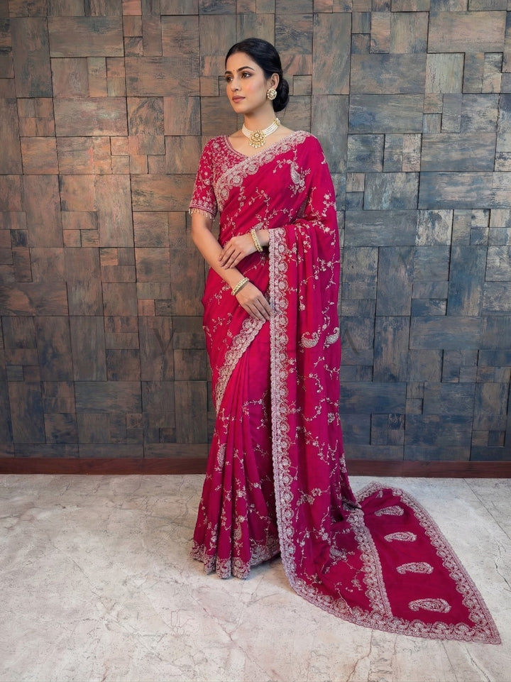 Woman in a pink saree with white patterns against a textured wall. Pink Satin Crepe Handwork Saree with Silver Embroidery Scalloped Bead &amp; Stone Work