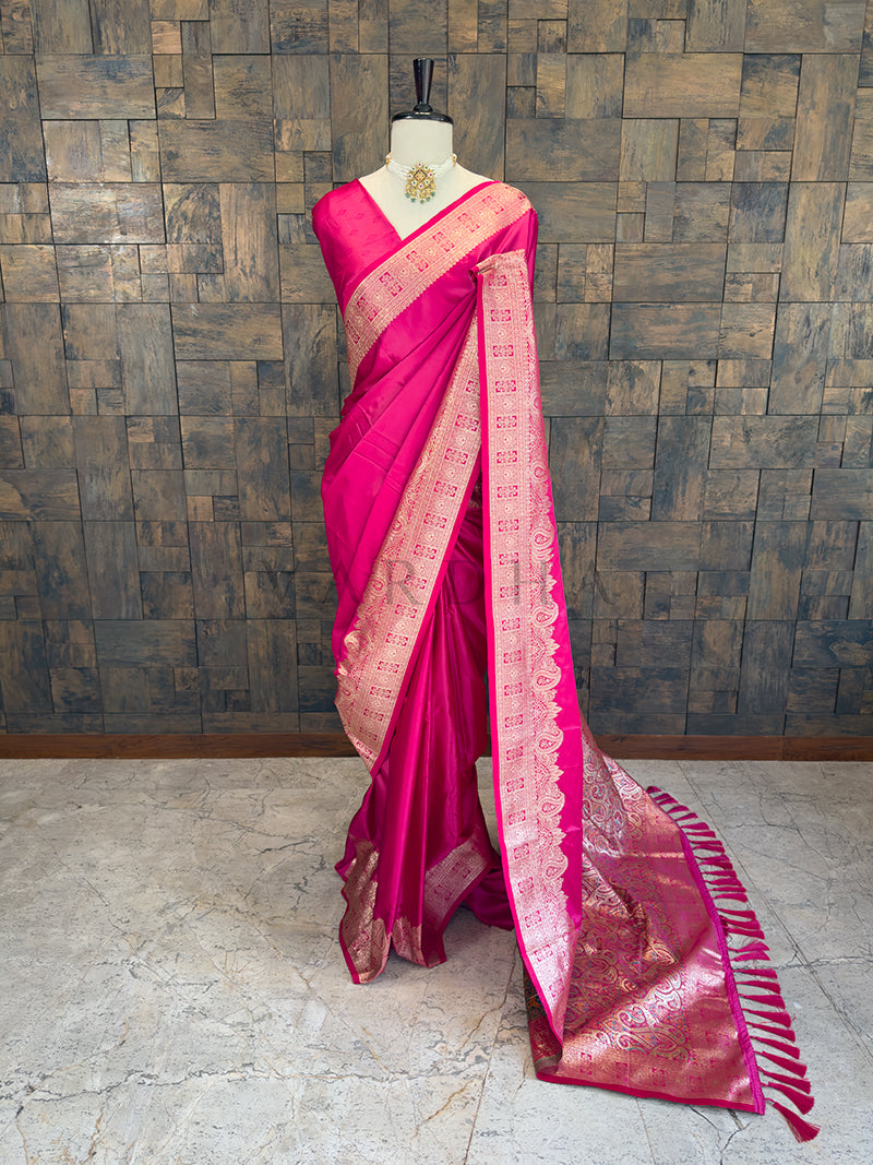 Pink saree with gold border on a mannequin against a textured wall. Full front display of a solid rani pink satin silk saree featuring a heavy golden zari border draped gracefully on a mannequin.