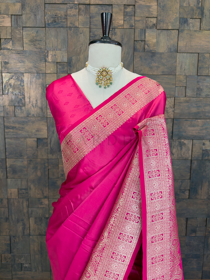 Pink saree with intricate patterns on a mannequin against a wooden paneled wall. Close-up of a bright pink woven satin ethnic saree styled on a mannequin display, paired with a traditional pearl choker necklace.