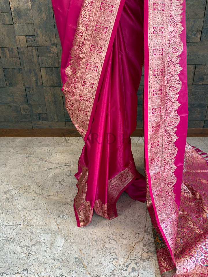 Pink saree with intricate patterns on a stone floor and wooden wall background Lower pleats and fluid drape of a glossy rani pink Banarasi-style satin silk saree showcasing the broad paisley border.