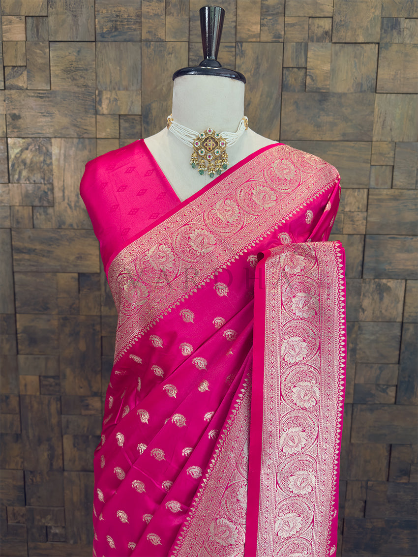 Pink saree with gold patterns on a mannequin against a textured wall background Close-up of a bright fuchsia woven satin ethnic saree styled on a mannequin display, paired with a traditional pearl choker necklace. Elegant party wear fuchsia satin silk saree highlighting its smooth drape, lustrous shine, and exceptional zari craftsmanship.