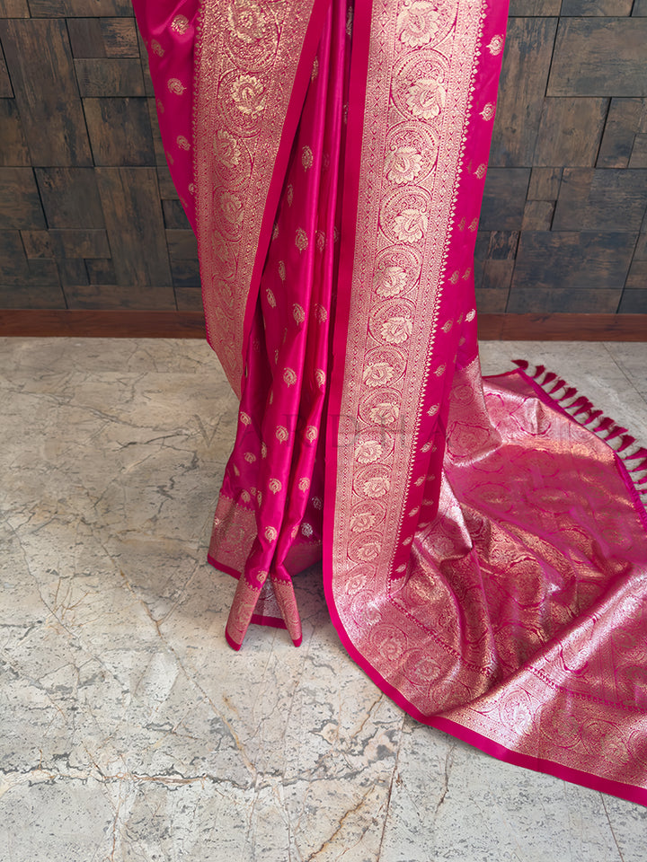 Pink saree with gold patterns on a marble floor and wooden wall Lower pleats and fluid drape of a glossy rani pink Banarasi-style satin silk saree showcasing the broad circular floral border.background
