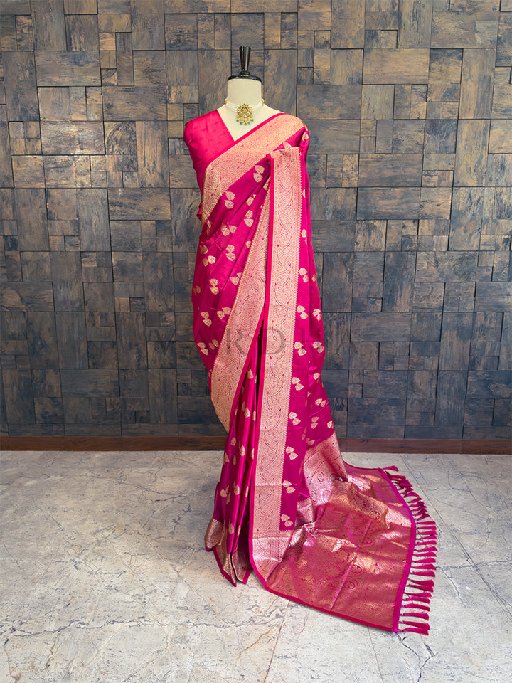 Pink saree with gold patterns on a mannequin against a wooden paneled wall. Full front display of a rani pink satin silk saree featuring golden zari bootis draped on a mannequin.