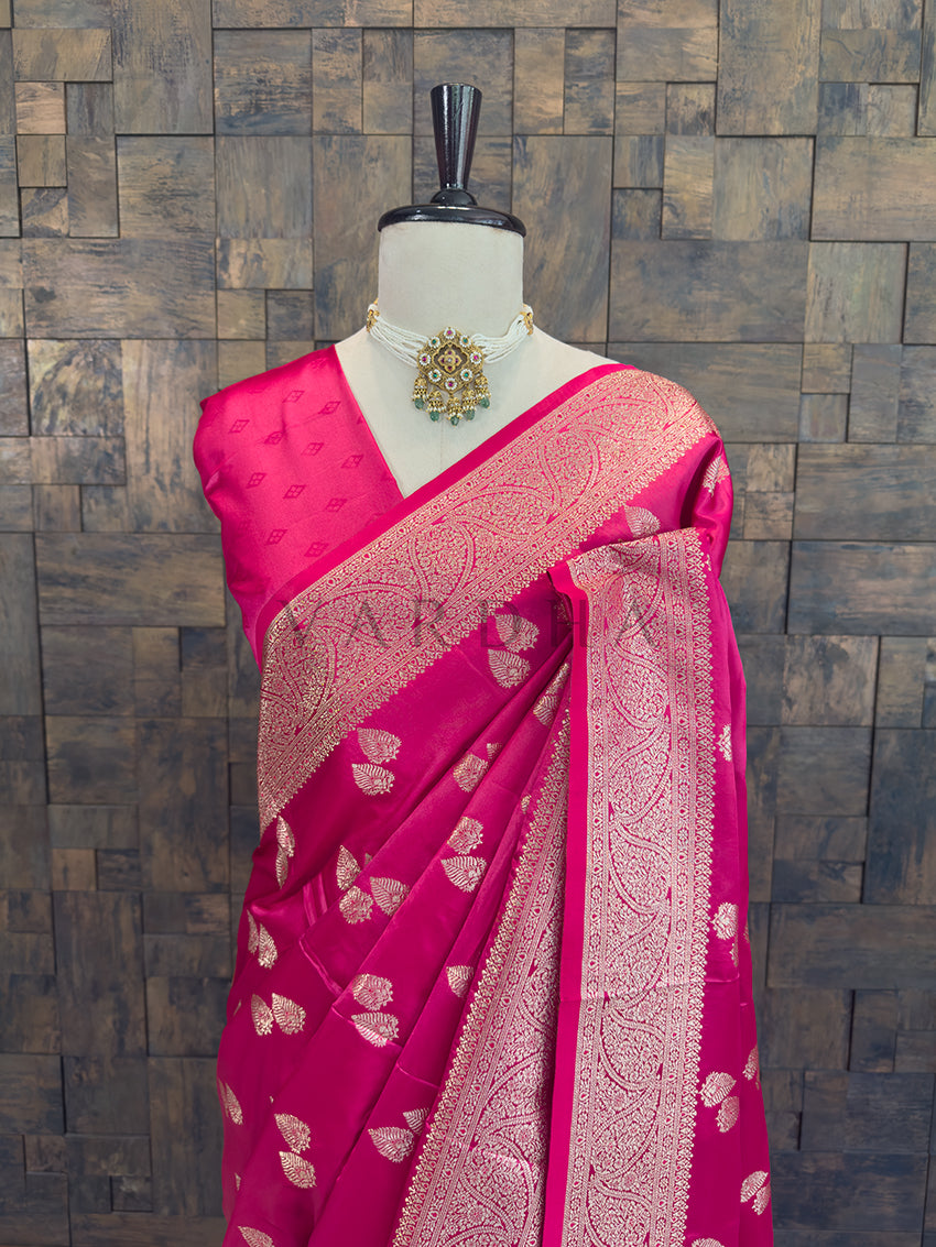 Pink saree with gold patterns on a mannequin against a wooden paneled wall. Close-up of the rani pink woven satin saree styled on a mannequin with a traditional pearl and emerald necklace.