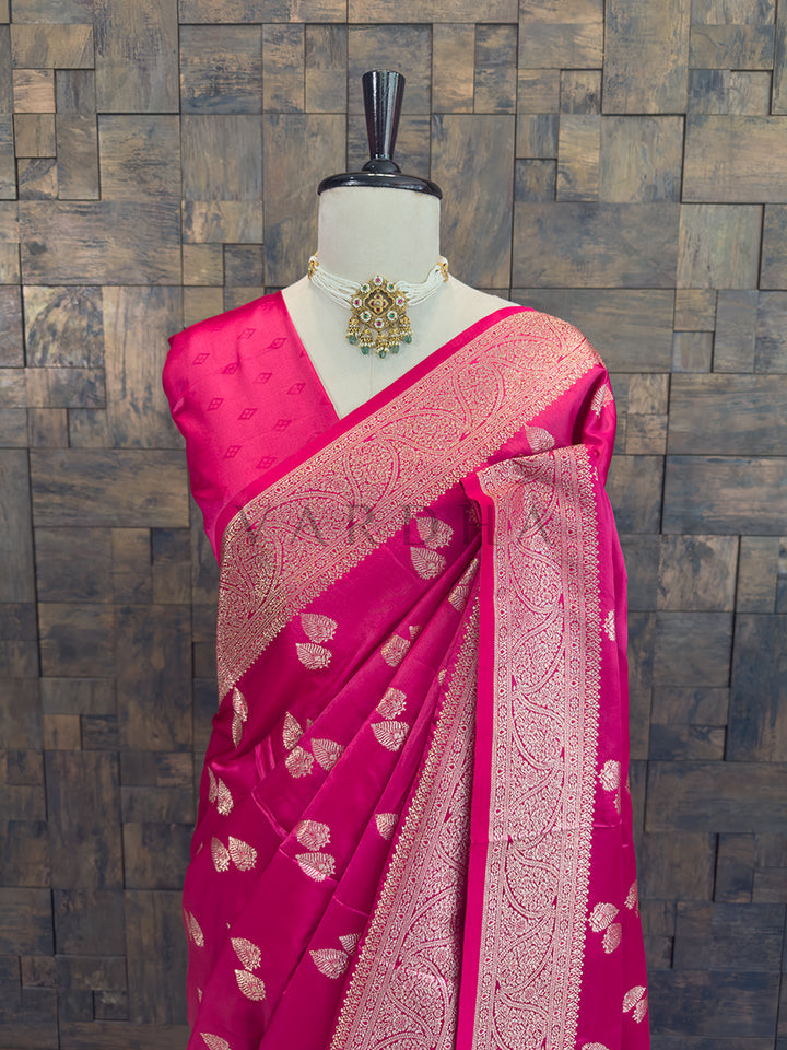 Pink saree with gold patterns on a mannequin against a wooden paneled wall. Close-up of the rani pink woven satin saree styled on a mannequin with a traditional pearl and emerald necklace.