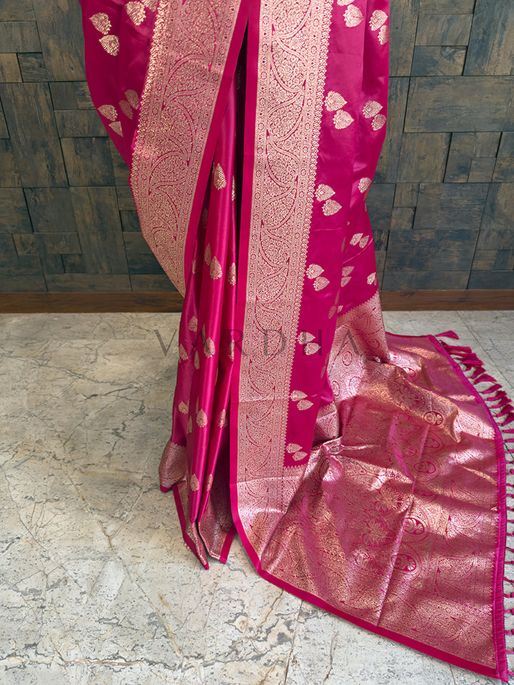 Pink saree with gold patterns on a stone floor and wooden wall background Detailed floor view of the rich golden zari woven pallu with matching pink tassels on a fuchsia satin saree. 