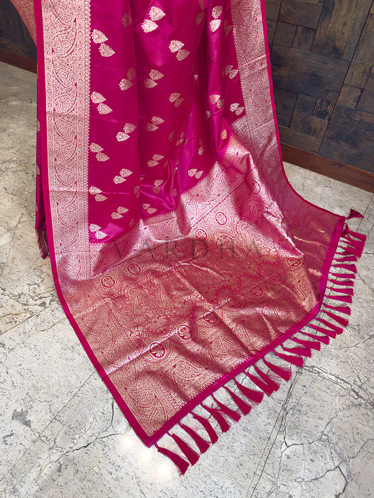 Pink saree with intricate patterns on a textured floor Close-up texture shot highlighting the golden leaf bootis and floral zari border weaving on pink satin fabric. Festive wear rani pink satin silk saree showcasing its glossy finish, fluid drape, and traditional metallic thread work.