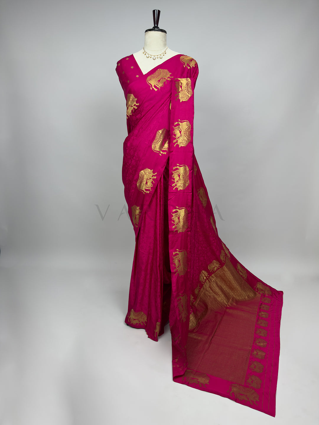 Pink saree with gold patterns on a mannequin against a gray background A full-length presentation of a vibrant Pink Satin Silk Saree beautifully draped on a mannequin against a neutral gray background. The saree features a bright rani pink body adorned with a subtle self-jacquard floral texture, dramatically contrasted by large, ornate gold zari elephant motifs woven across the drape. The heavy, gold-woven pallu flows elegantly onto the floor, crafting a luxurious, eye-catching ensemble perfect for weddings