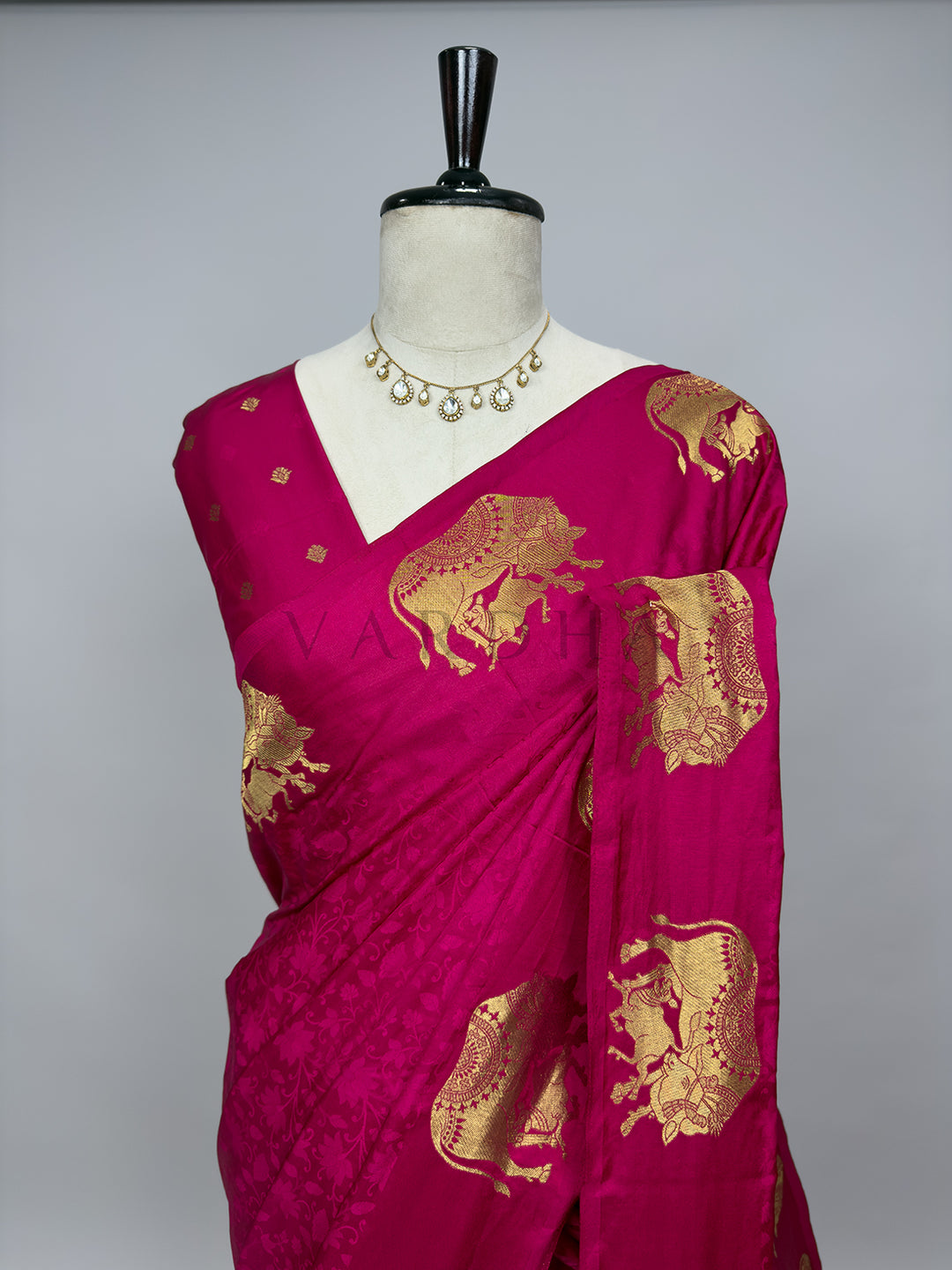 Pink saree with gold patterns on a mannequin against a gray background A detailed half-torso perspective of the draped Pink Satin Silk Saree on a mannequin, styled with a classic stone statement necklace. This angle highlights the exquisite pattern layout over the chest and pleated shoulder drape, emphasizing the embossed tone-on-tone floral vines woven into the glossy pink silk base alongside the intricately detailed, shimmering gold zari elephant motifs.