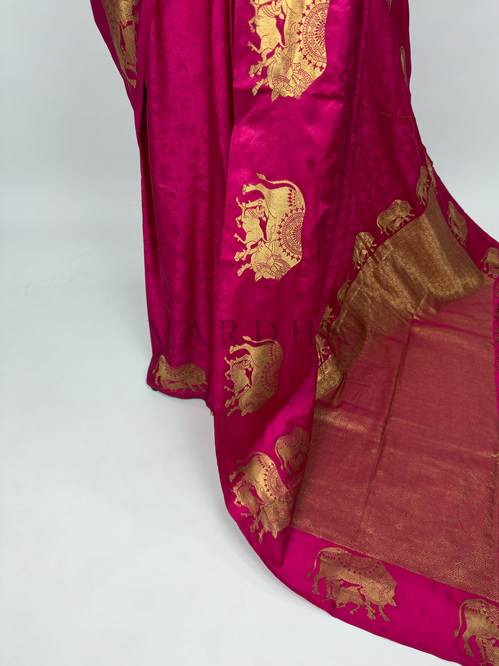 Pink saree with gold embroidery on a white background A targeted shot focusing on the lower drape and front pleats of the Pink Satin Silk Saree. The image showcases the smooth, reflective sheen of the premium satin fabric as it forms crisp, structured vertical folds. A prominent vertical panel decorated with a repeating sequence of gold zari elephant motifs runs down the side, emphasizing a clean silhouette and leading into the grand pallu.
