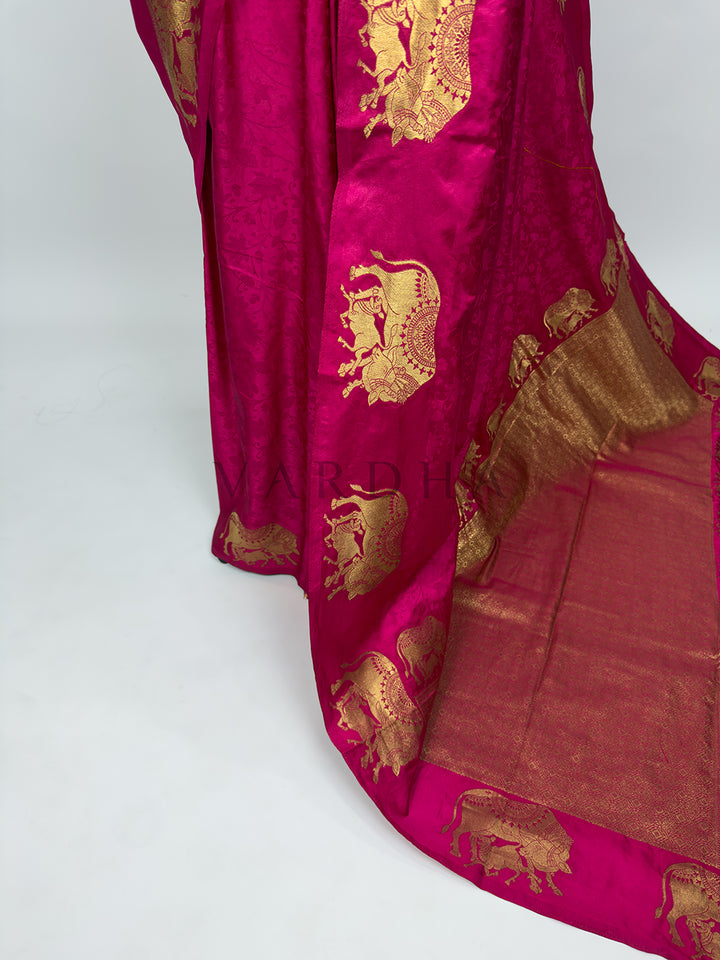 Pink saree with gold embroidery on a white background A targeted shot focusing on the lower drape and front pleats of the Pink Satin Silk Saree. The image showcases the smooth, reflective sheen of the premium satin fabric as it forms crisp, structured vertical folds. A prominent vertical panel decorated with a repeating sequence of gold zari elephant motifs runs down the side, emphasizing a clean silhouette and leading into the grand pallu.
