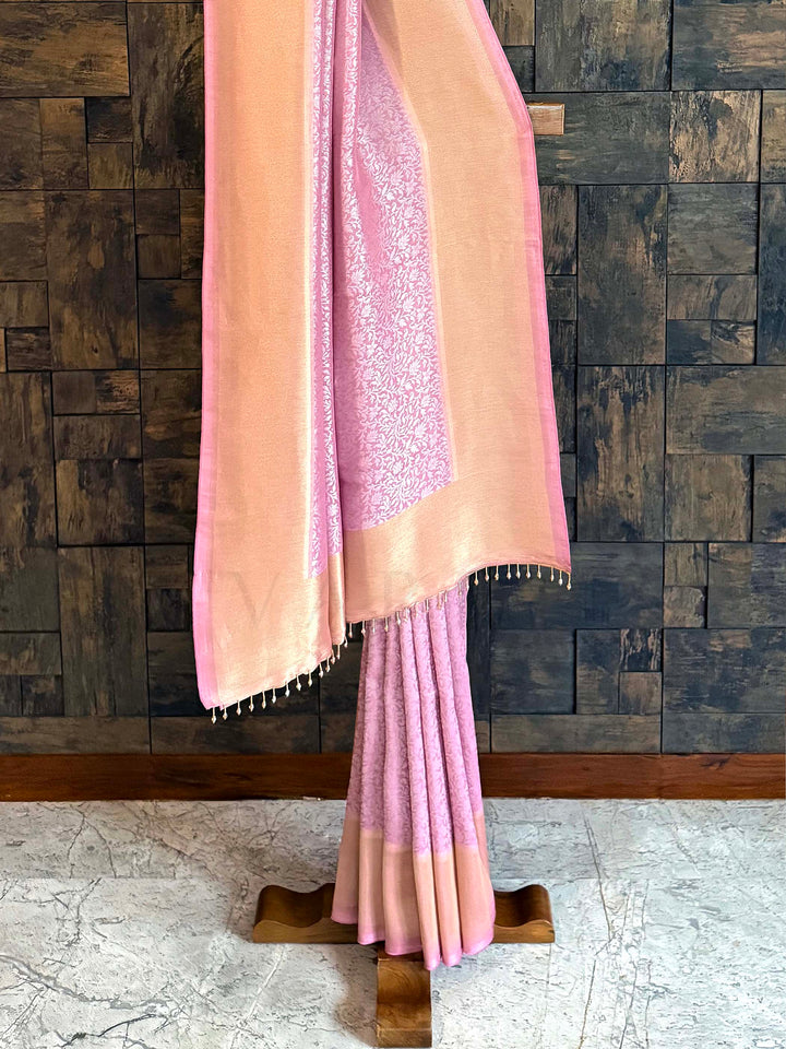 Pink saree with a textured border draped over a wooden rod against a wooden paneled wall. Pallu shot featuring rose panel framed in peach-gold tissue with tassel edging.