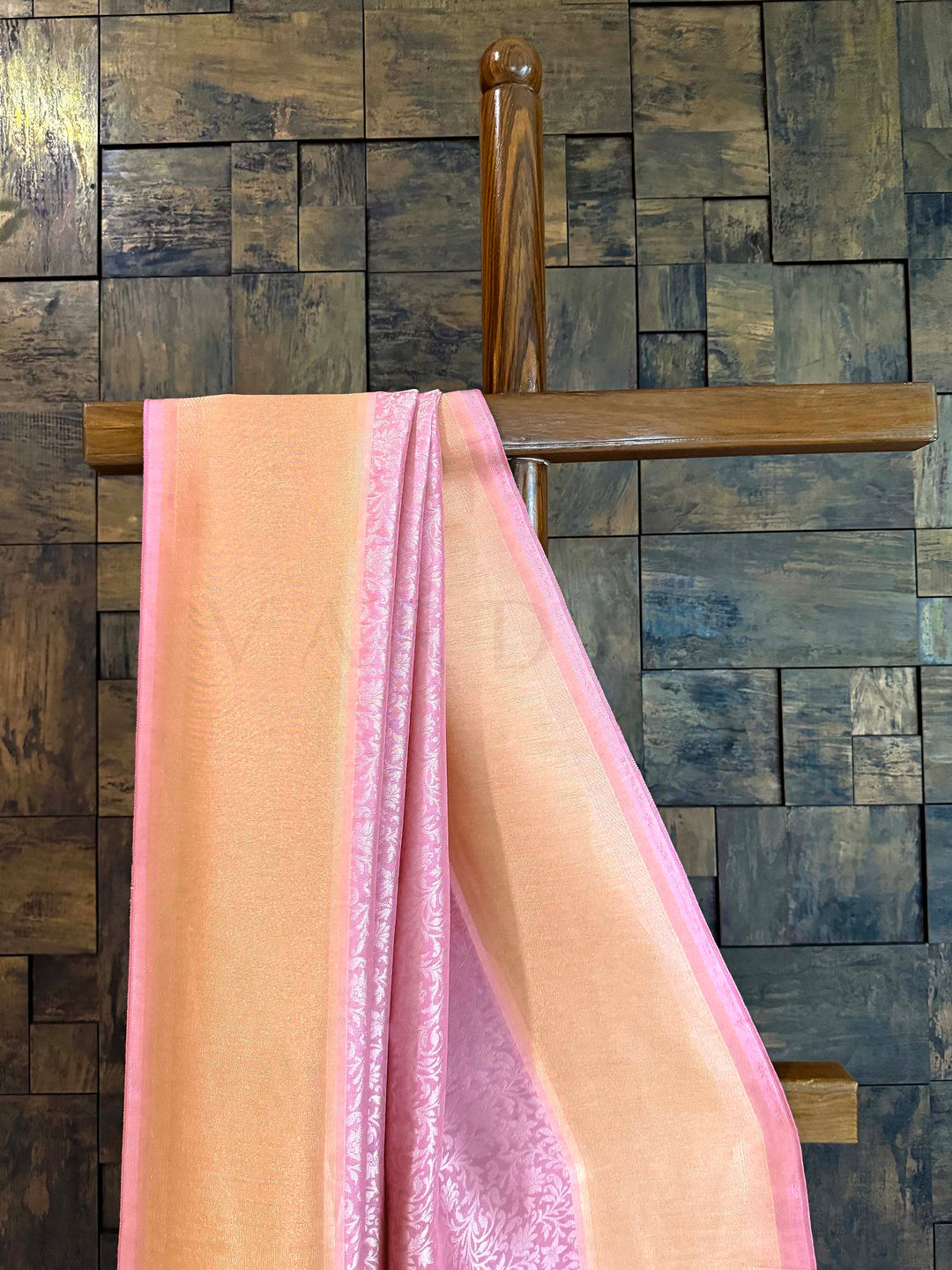 Pink fabric draped over a wooden rod against a wooden paneled wall. Pleated section of saree showing fine floral jaal and luminous border. Edge view capturing tasselled finish and refined zari tissue frame.