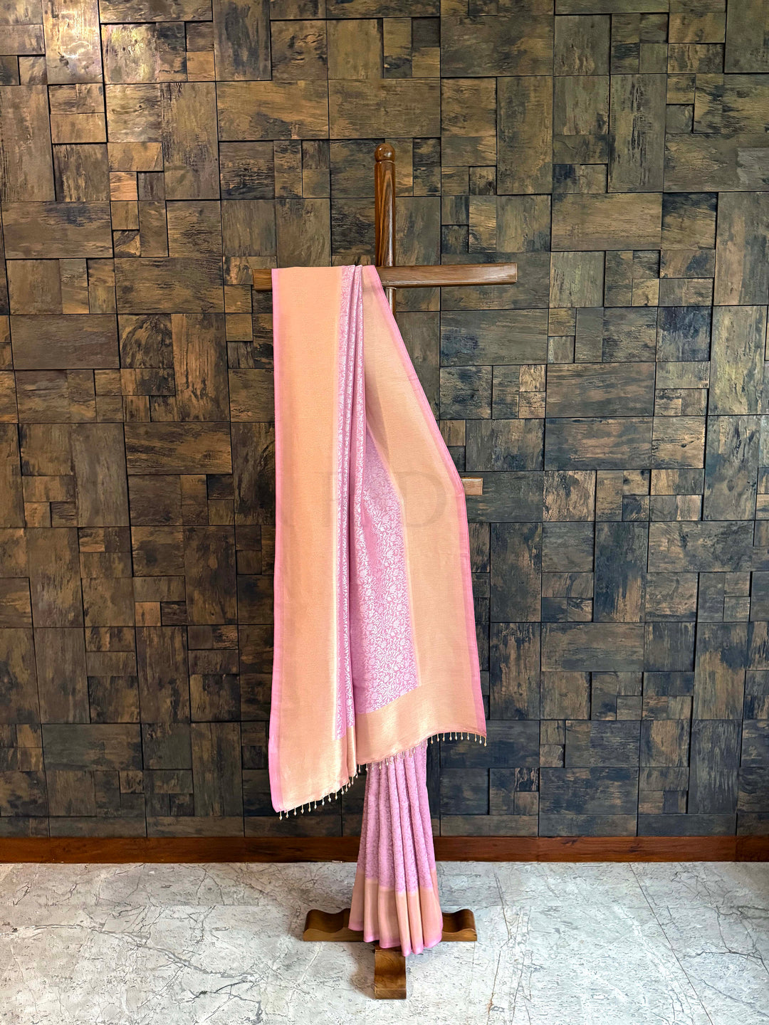 Full-length display of pink–peach Kanjivaram Tissue Silk saree highlighting two-tone glow. Pink saree with a wooden hanger against a textured wall. Coordinating blouse piece placed with the saree, matching border and soft sheen. Detailed view of woven floral creeper jaal on rose-pink tissue silk fabric.