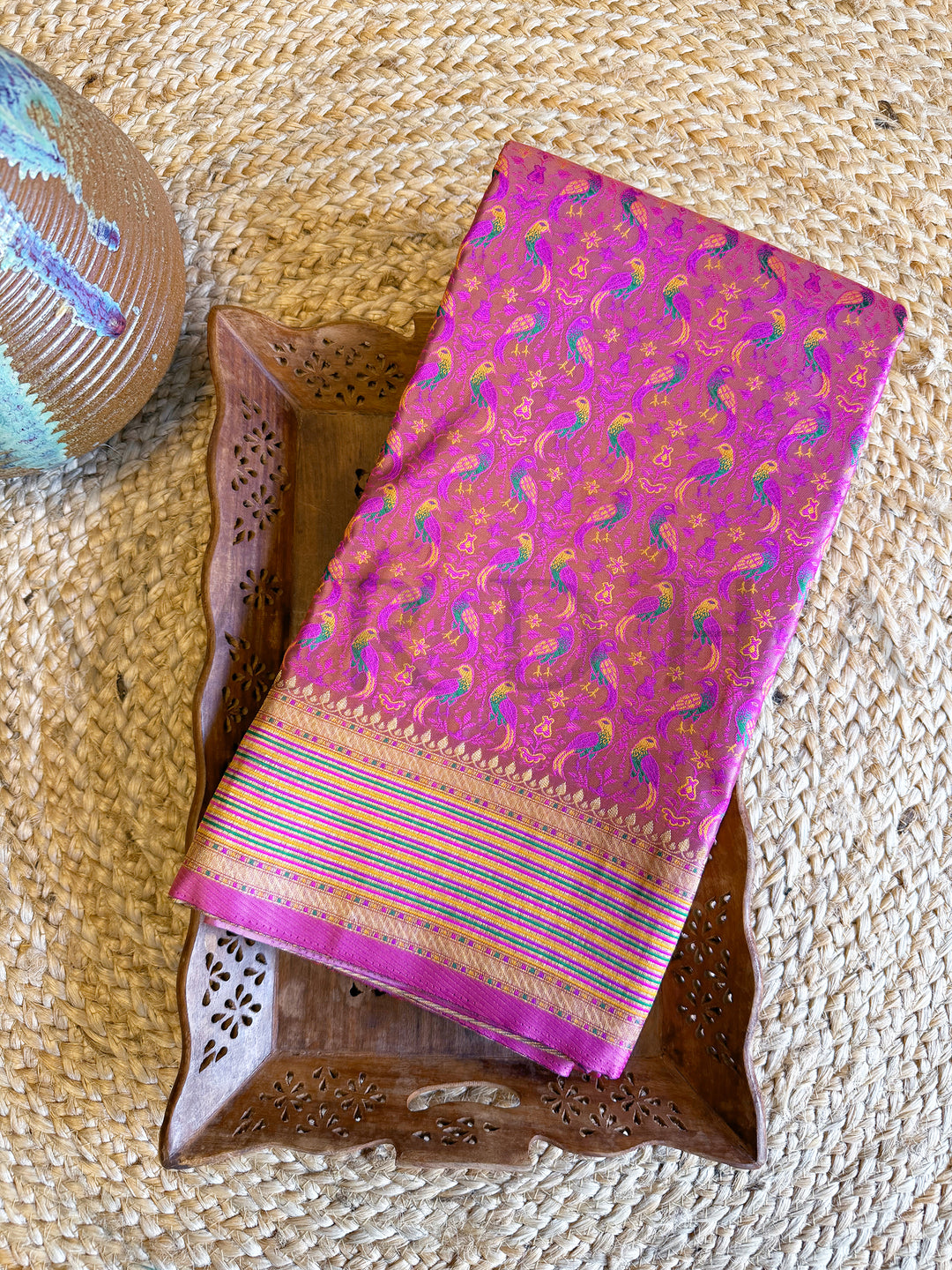 Folded pink satin silk saree with thread-woven parrot motifs and gold zari border, displayed on rustic tray for product showcase