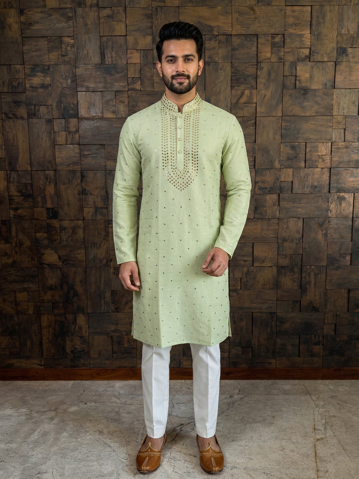 Man wearing a light green kurta with white pants against a wooden paneled wall. Pista green khadi cotton kurta with gold mukaish work styled with white churidar and brown mojari