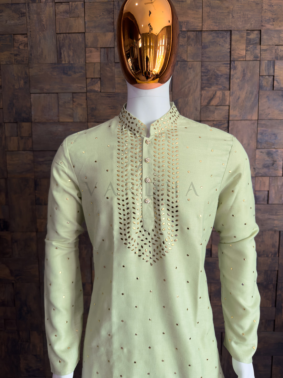 Light green kurta with intricate embroidery on a mannequin against a wooden paneled wall. Traditional pista green khadi kurta with subtle gold mukaish detailing perfect for wedding functions