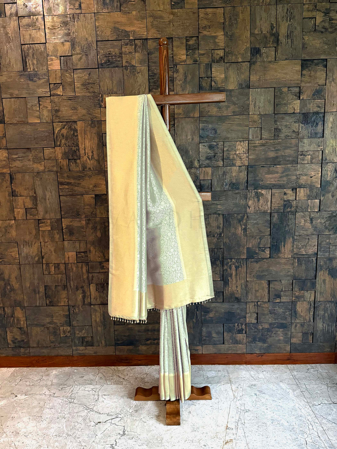 Beige and white saree draped over a wooden hanger against a wooden paneled wall. Pista Green Icy mint and butter-gold Kanjivaram Tissue Silk saree with floral jaal displayed on a wooden stand. 