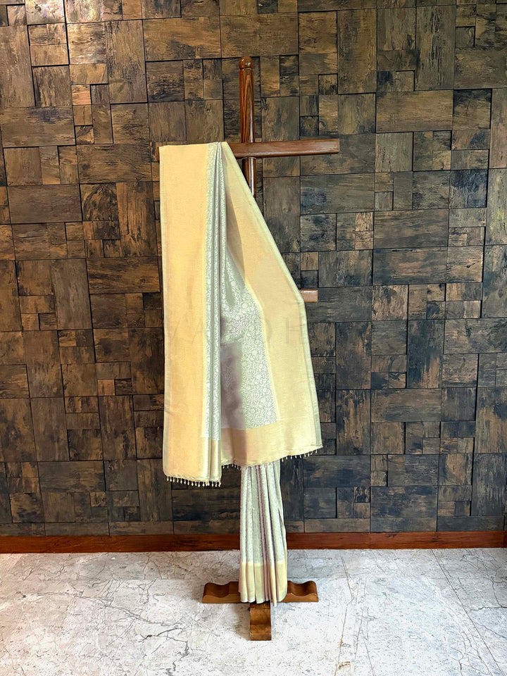Beige and white saree draped over a wooden hanger against a wooden paneled wall. Pista Green Icy mint and butter-gold Kanjivaram Tissue Silk saree with floral jaal displayed on a wooden stand. 