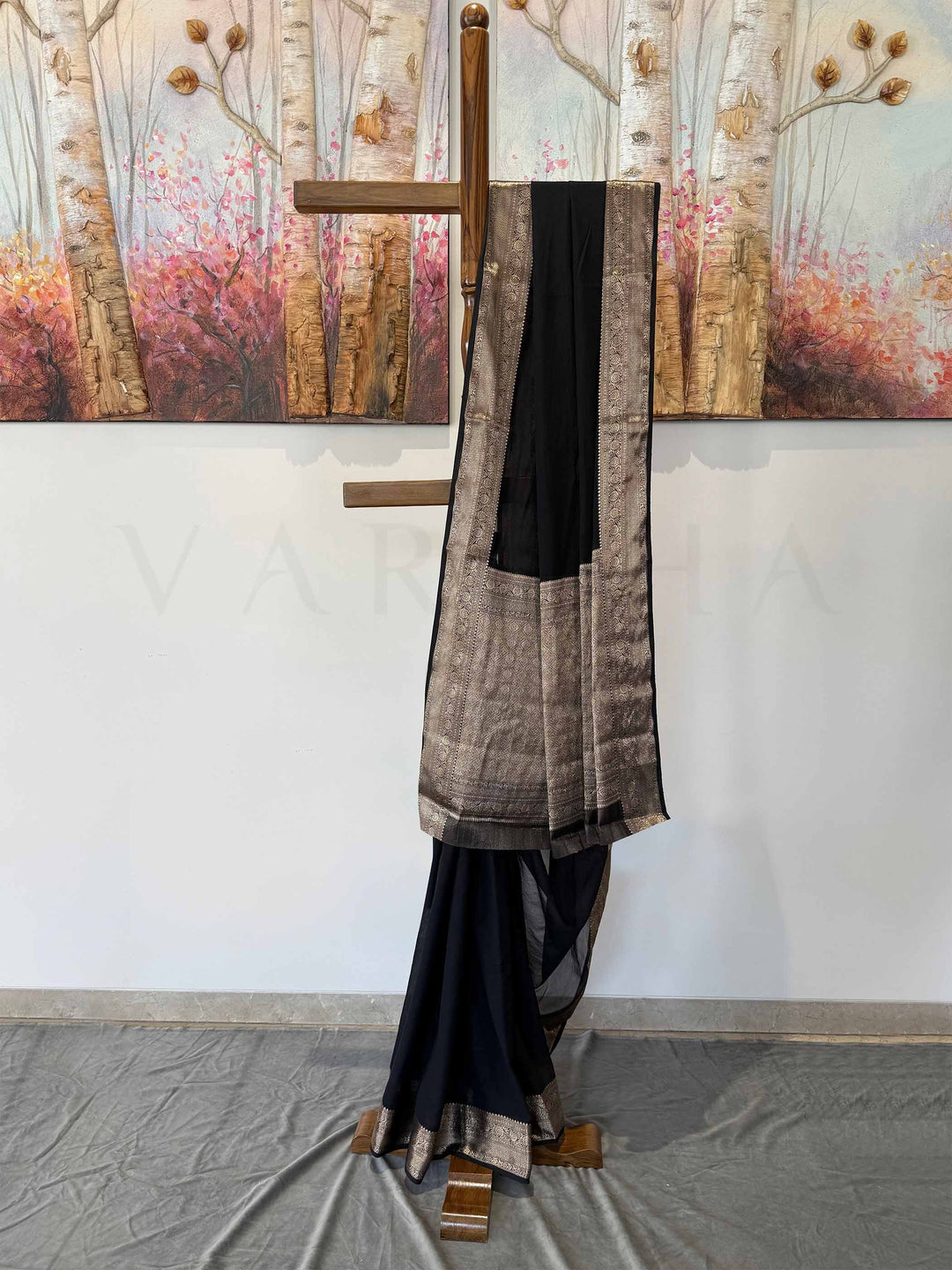 A stunning black georgette saree with gold zari work, gracefully displayed on a wooden stand. The delicate weave and intricate border detailing highlight its luxurious appeal, perfect for special occasions.