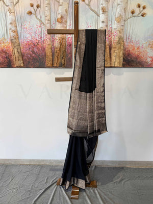 A stunning black georgette saree with gold zari work, gracefully displayed on a wooden stand. The delicate weave and intricate border detailing highlight its luxurious appeal, perfect for special occasions.