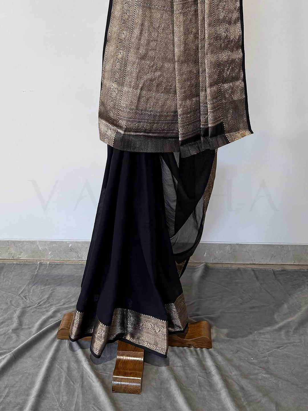 An exquisite black georgette saree with a wide gold zari border, carefully draped on a wooden frame. The soft, flowing fabric reflects sophistication, making it an ideal choice for festive and formal wear.