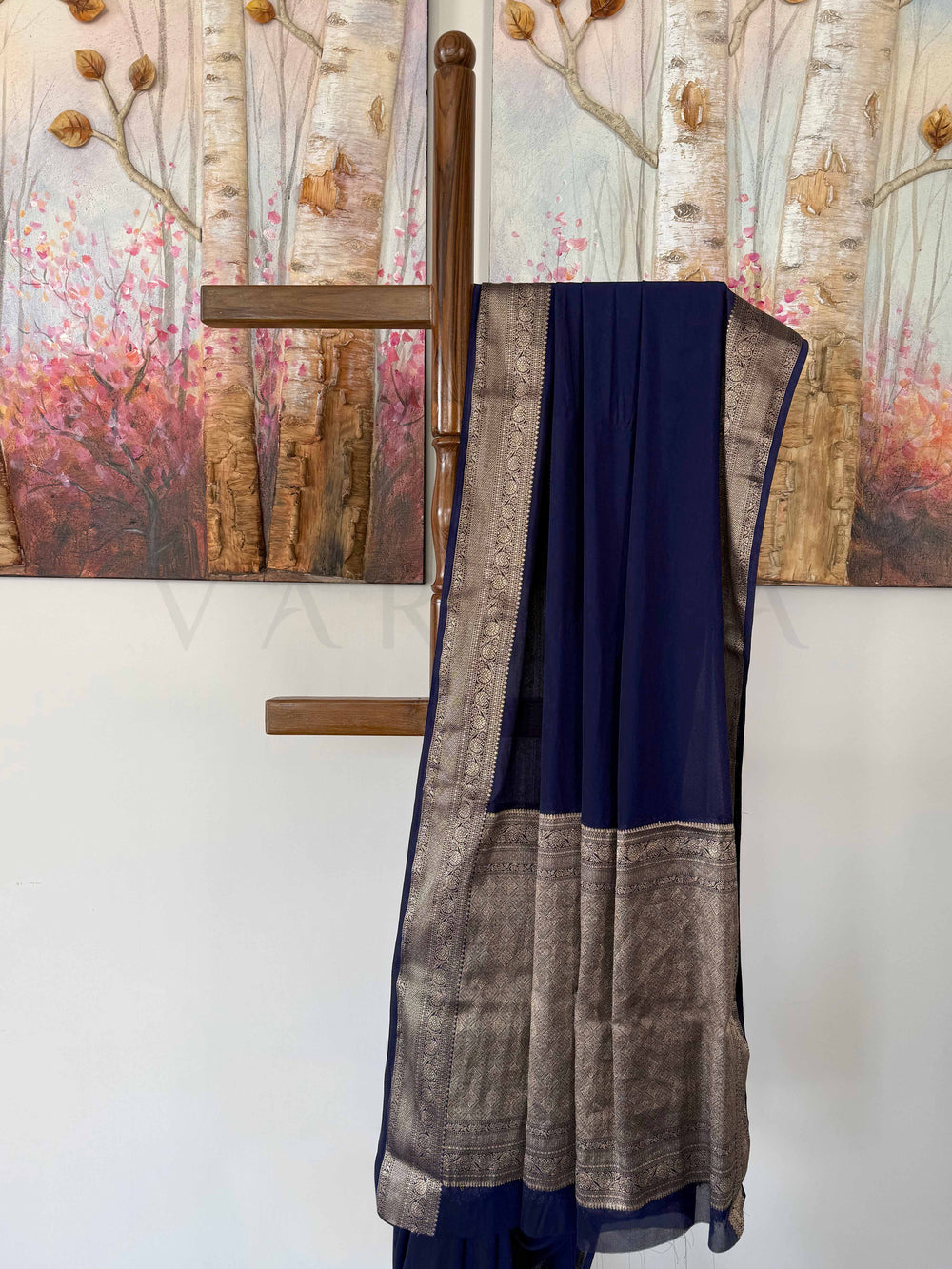 A luxurious deep blue georgette saree with intricate gold zari work, gracefully displayed against a warm artistic background. The saree’s elegant drape and timeless craftsmanship make it a standout piece.
