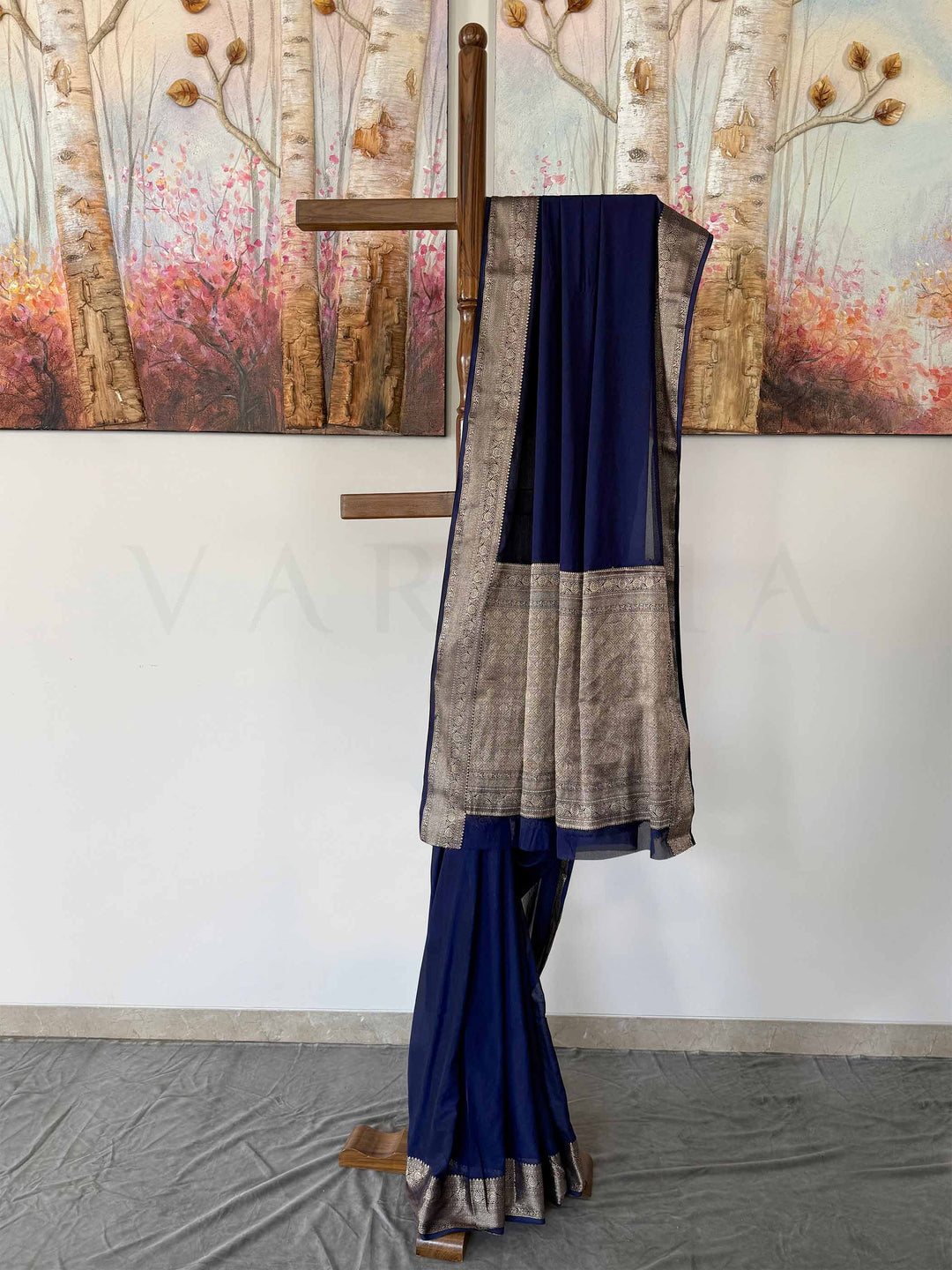 A stunning navy blue georgette saree with shimmering gold zari embroidery, elegantly arranged on a wooden stand. The soft, flowing fabric and intricate detailing add to its rich, traditional charm.