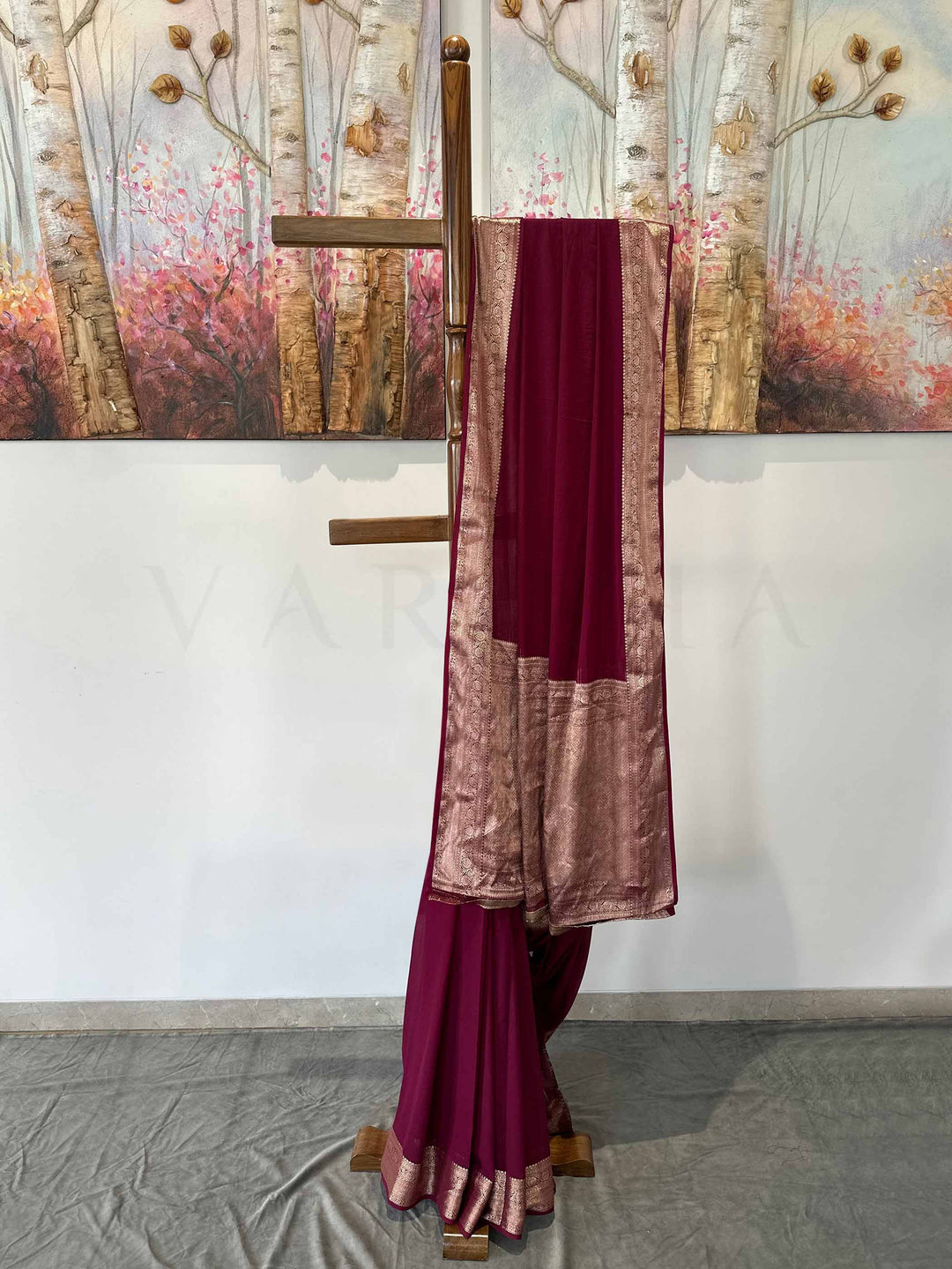 A luxurious wine georgette saree with delicate gold zari work, beautifully displayed on a wooden stand. The soft, flowing fabric drapes effortlessly, showcasing its refined craftsmanship and timeless appeal