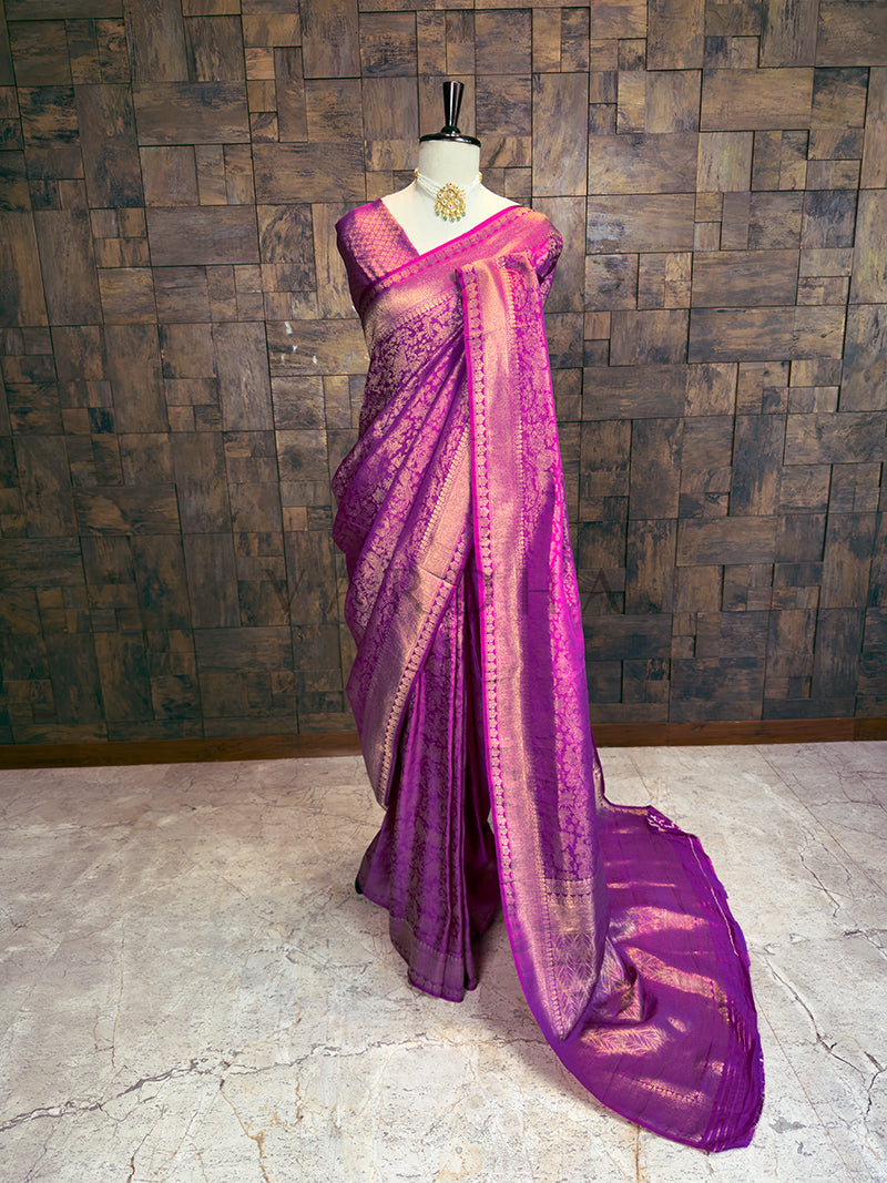 Full-length draped view of a stunning Purple Kanjivaram Silk Saree by House of Vardha, highlighting the contrast between the rich purple silk and lustrous gold zari weave. We offer Ready to wear, Pre drape service, Saree Add on, Custom Stitched Blouse, Fall and Edging, and Gift Packaging at Complimentary Price!. Purple saree with pink border on a mannequin against a wooden paneled wall.