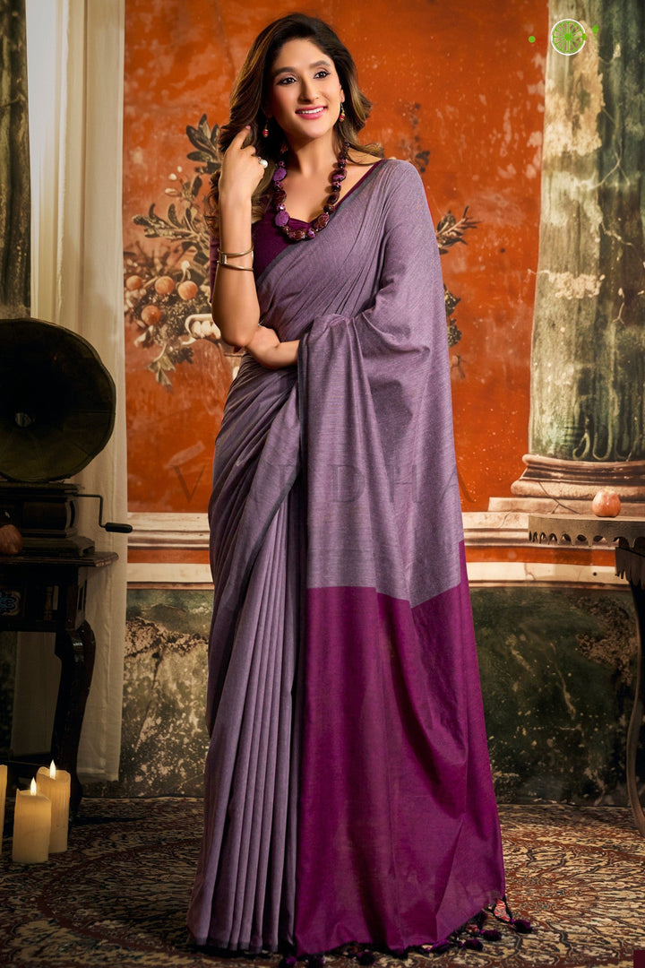 A woman models a two-tone purple saree with a solid lavender upper drape and a deep magenta lower pallu, paired with a matching purple blouse, set against an ornate vintage interior background. Full front view of a woman wearing a comfortable purple mal cotton saree featuring a dark magenta contrast pallu with fancy tassels.