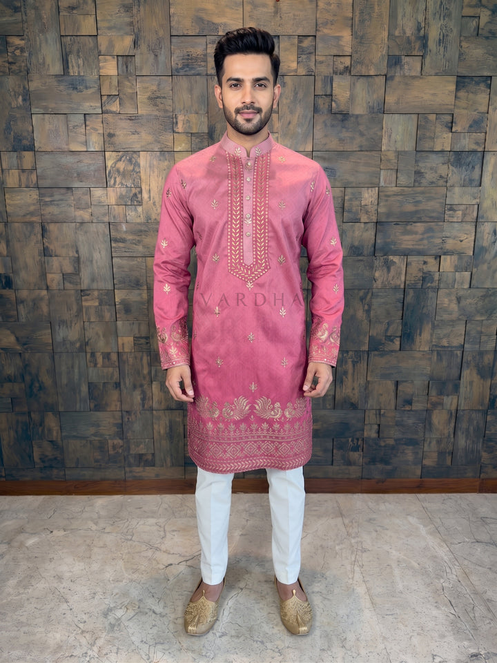 Man wearing a pink embroidered kurta with white pants against a wooden paneled wall.  kurta boasts a subtle tonal self-pattern that adds depth and a premium sheen. The kurta features a striking heavy designer yoke, intricately embroidered with gold leafy vines. 