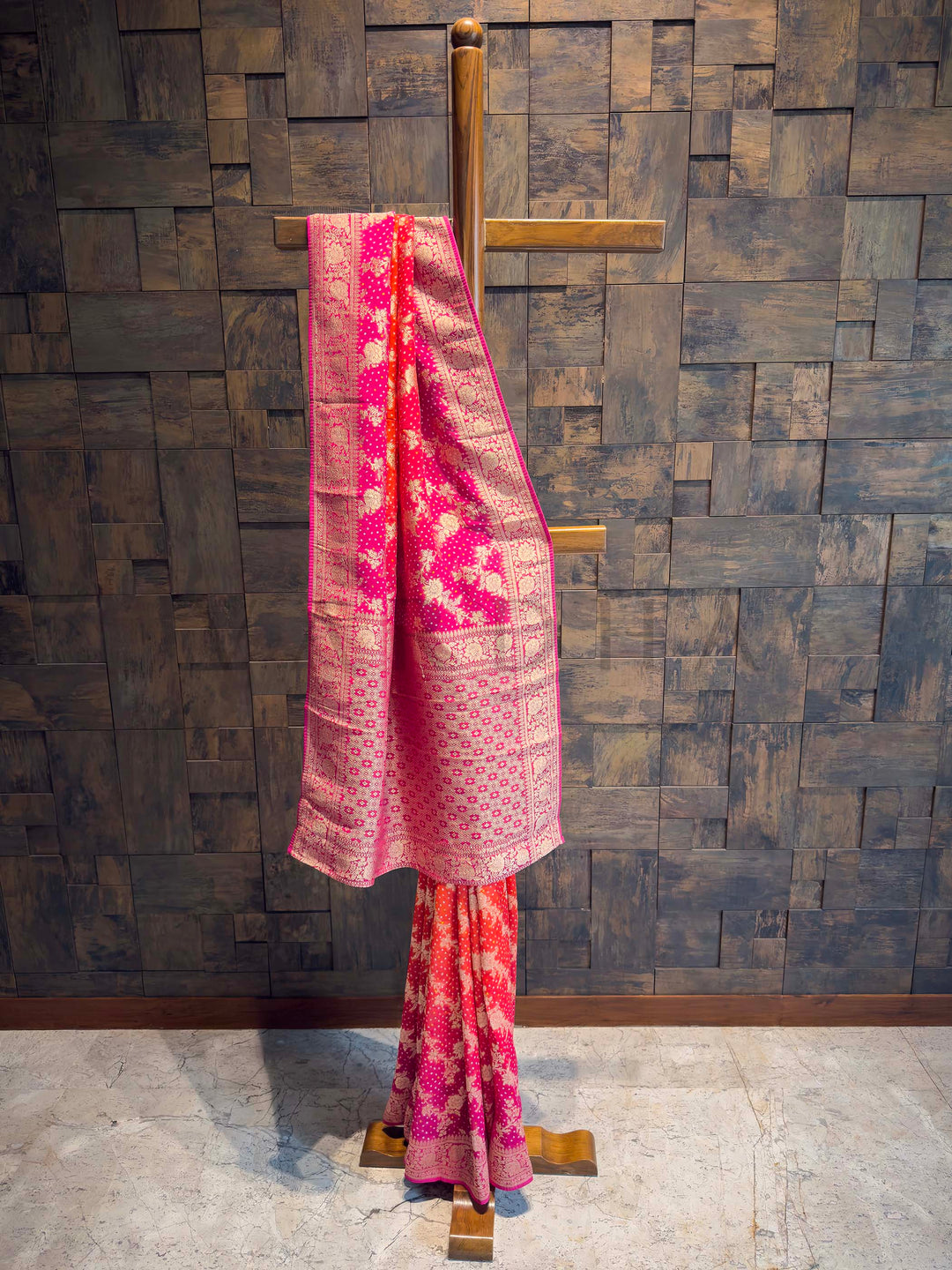 Pink saree with floral patterns draped over a wooden rod against a textured wall.
