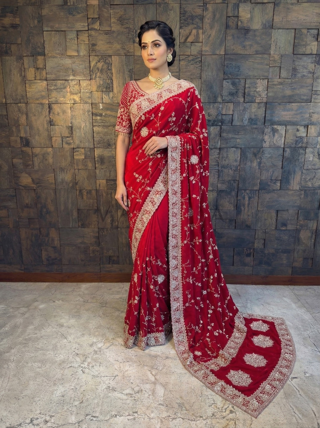 Woman wearing a red saree with a textured wall background. Red Satin Crepe Saree with Silver Cutdana & Floral Zircon Handwork