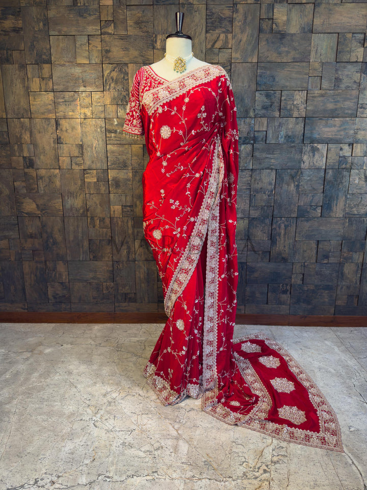 Red saree with white patterns on a mannequin against a textured wall. House of Vardha Red Satin Crepe Saree featuring Silver Cutdana &amp; Floral Zircon Handwork, elegantly draped on a mannequin. We offer Ready to wear, Pre drape service, Saree Add on, Custom Stitched Blouse, Fall and Edging, Gift Packaging at Complimentary Price!