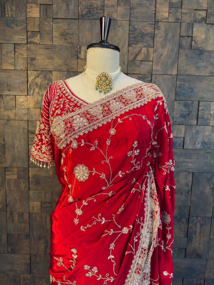 Red saree with white embroidery on a mannequin against a wooden paneled wall. Elegant Red Satin Crepe Saree by House of Vardha showcasing exquisite Silver Cutdana &amp; Floral Zircon Handwork alongside a matching heavily embellished blouse. We offer Ready to wear, Pre drape service, Saree Add on, Custom Stitched Blouse, Fall and Edging, Gift Packaging at Complimentary Price!