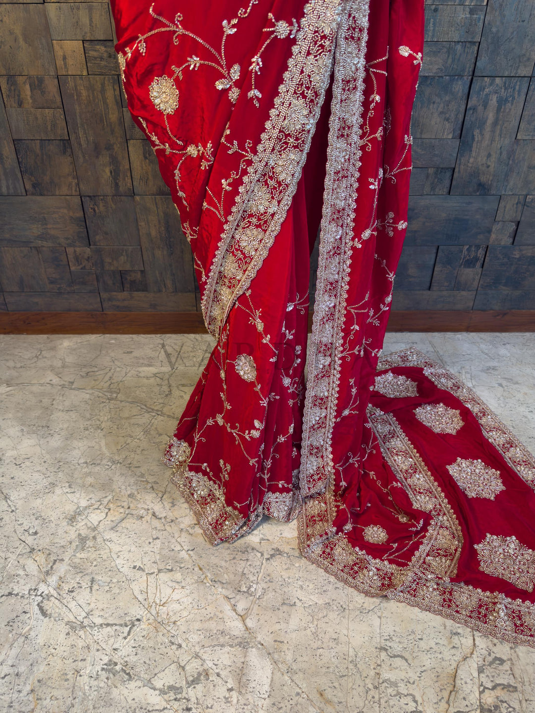 Red embroidered saree with gold patterns on a stone floor and wooden wall background. Close-up view of the House of Vardha Red Satin Crepe Saree highlighting the detailed Silver Cutdana &amp; Floral Zircon Handwork and delicate scalloped border. We offer Ready to wear, Pre drape service, Saree Add on, Custom Stitched Blouse, Fall and Edging, Gift Packaging at Complimentary Price!