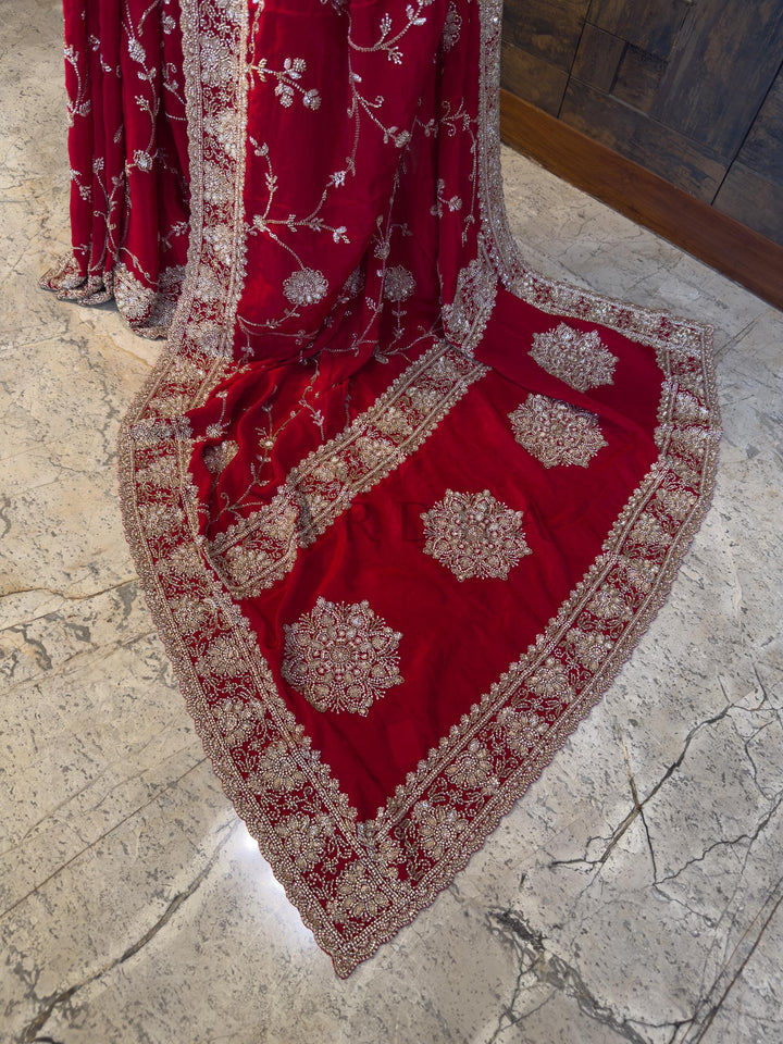 Red embroidered fabric on a stone floor. Luxurious Red festive Satin Crepe Saree from House of Vardha featuring radiant Silver Cutdana &amp; Floral Zircon Handwork, perfect for glamorous evening parties and weddings. We offer Ready to wear, Pre drape service, Saree Add on, Custom Stitched Blouse, Fall and Edging, Gift Packaging at Complimentary Price!