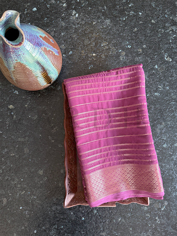 Folded rose pink lightweight georgette saree featuring golden zari border and floral pallu, displayed with vase and tray