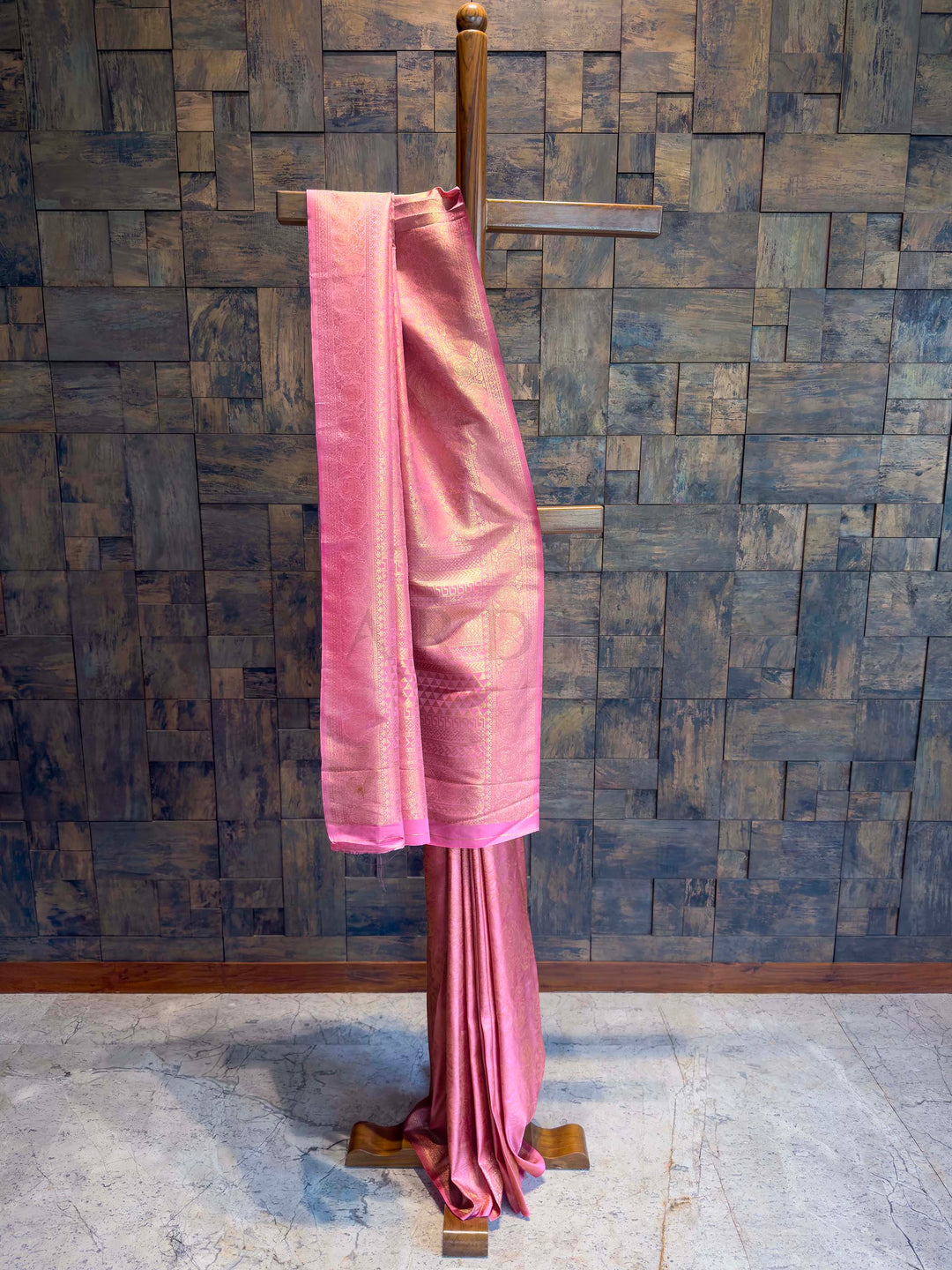 Pink saree with gold border on a wooden stand against a textured wall. Rose Pink Kanjivaram Silk Saree with intricate golden zari weaving displayed on hanger