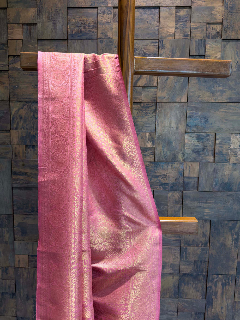 Pink fabric draped over a wooden rod against a textured blue wall. Close-up view of rose pink saree border featuring circular golden zari motifs